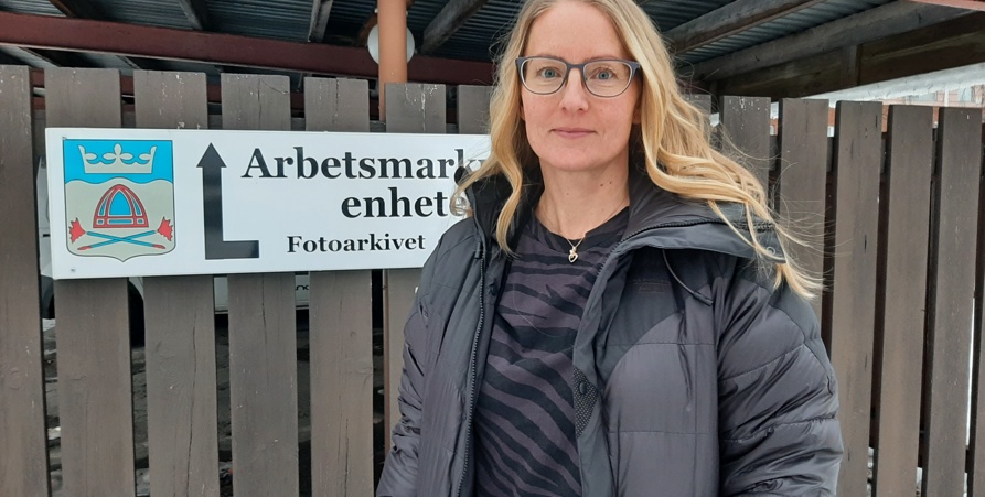 Ann-Eva Holmström fotograferad utomhus framför en skylt med Vilhelmina kommuns logotyp och texten Arbetsmarknadsenheten. 
