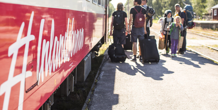 Bilden visar sidan av en tågvagn med texten "#Inlandsbanan". På perrongen står en grupp människor med ryggen mot kameran redo att gå på tåget. 