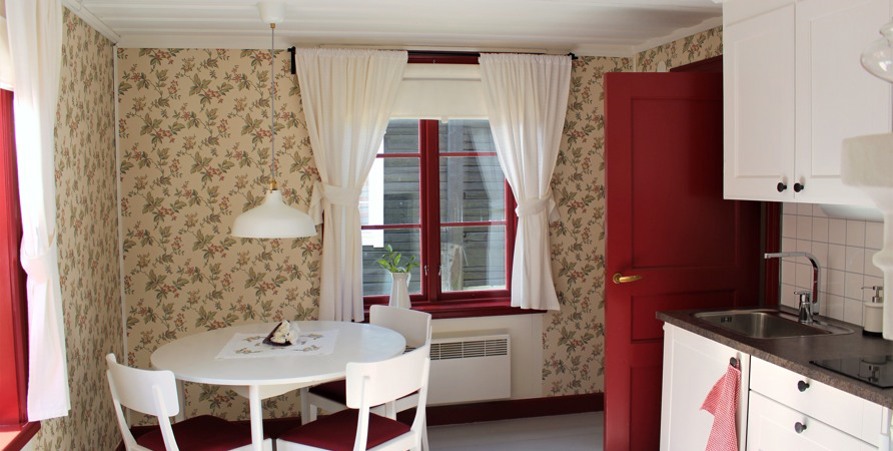 Interiör från ett av rummen i ett av husen i Kyrkstan. Ett rum med mönstrade tapeter med ett vitt bord som står vid ett fönster med vita gardiner. 