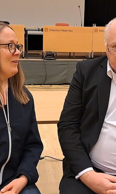 Två personer sitter ner. Den till vänster tittar på den till höger som tittar in i kameran.