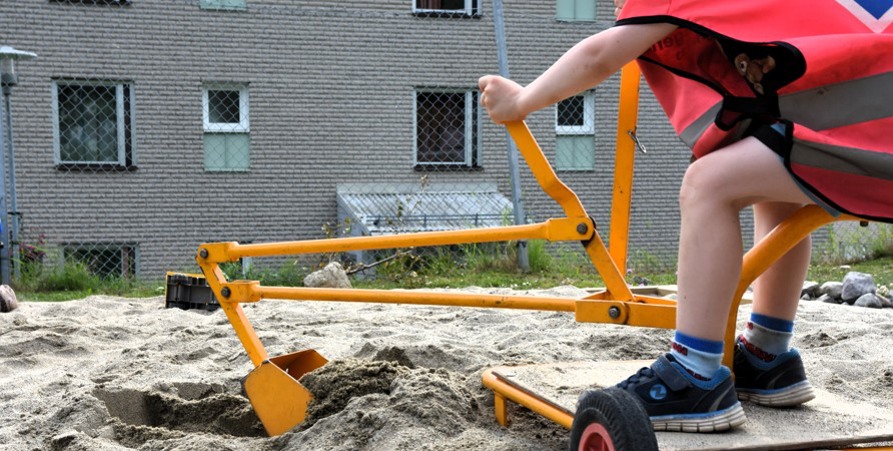En bild där man ser ett barns armar och ben som styr en lekgrävskopa i sandlådan. 