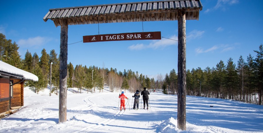 Bilden visar starten vid skidstadion. Tre personer på längdskidor åker under skylten "I Tages spår". 